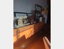 Eine hölzerne Anrichte in einem Raum, dekoriert mit gerahmten Bildern, Metallskulpturen, einer Wanduhr, einem mehrarmigen Kerzenleuchter und einer Tischlampe. Sonnenlicht fällt durch ein Fenster und wirft Schatten auf die Möbel und den Boden.