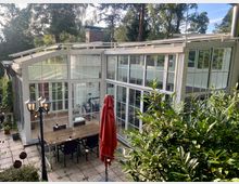 Modernes Wintergarten-Gebäude mit großen, weißen Glasfassaden und einem flachen Satteldach, das in einer begrünten Umgebung steht. Im Vordergrund befindet sich eine Terrasse mit einem Holztisch, Stühlen, einem roten Sonnenschirm und einer Laterne.