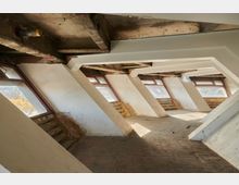 Loftartige Innenansicht eines unrenovierten Dachgeschosses mit großen, schräg angebrachten Fenstern und freiliegenden Holzbalken. Der Raum ist leer und weist deutliche Gebrauchsspuren an Wänden und Boden auf.