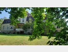 Großes, mehrstöckiges Landhaus mit hellgelber Fassade und dunklem Schrägdach, umgeben von einem gepflegten Garten. Der Blick durch die grünen Baumblätter zeigt eine Terrasse mit Treppe und einen kleinen Baum mit blühenden Ästen.
