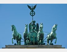 Bronzefigur einer Quadriga mit vier Pferden, gelenkt von einer geflügelten Frauengestalt, die einen Stab mit Eichenkranz und Adler hält, auf einem Gebäudegiebel vor klarem, blauem Himmel.