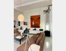 Modern living room with white walls and high ceiling beams, featuring a brown sectional sofa facing a low black coffee table on a light rug. The space includes a large orange-toned painting above sleek white cabinetry, pendant and floor lamps, built-in shelving, and a sculptural branch-like ceiling fixture.