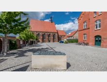 Pflastersteinplatz mit Bäumen und Sitzbänken vor einer historischen Backsteinkirche mit großen gotischen Fenstern und roten Ziegeldächern. Rechts im Bild stehen ein modernes Backsteingebäude mit Fahrrädern und weitere Häuser mit roten Dächern.