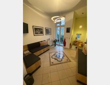 Heller Wartebereich mit Fliesenboden und einem dekorativen Muster in der Mitte, einer Sitzbank mit schwarzer Polsterung, an den Wänden anhängenden Fotos, einer großen Glastür mit Blick ins Grüne und einer modernen, kreisförmigen Deckenleuchte.