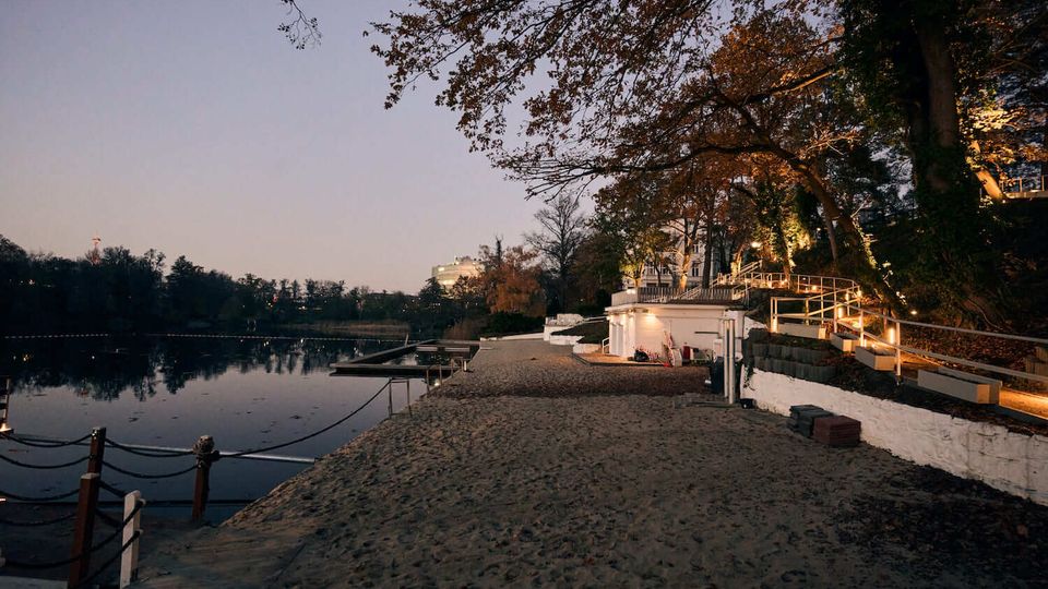 Lakeside waterfront at dusk with a sandy shoreline and calm water, bordered by small docks and rope railings. A terraced walkway with white retaining walls and wooden handrails runs along a tree-covered slope with warm lights and buildings set back above the shore.