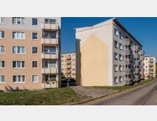 Mehrstöckige Wohnblöcke in einer Wohnsiedlung, mit hellen, pastellfarbenen Fassaden und Balkonen. Im Vordergrund ein gepflasterter Gehweg und eine Grünfläche; im Hintergrund weitere ähnliche Gebäude und geparkte Autos.