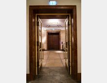 Doppeltür aus Holz mit Glaseinsätzen und goldfarbenen Griffen, eingebettet in einen braunen Marmorahmen, führt zu einem Innenraum mit beige gefliesten Wänden und Böden. Ein Notausgangsschild leuchtet über der Tür.