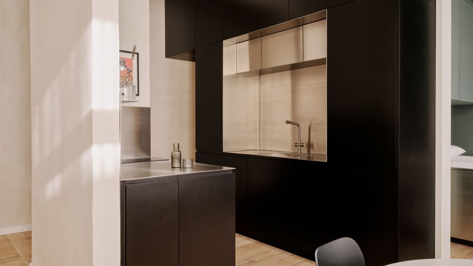 Moderne, minimalistische Küche mit mattschwarzen Einbauschränken und einer großen Nische aus Edelstahl mit Spüle und Armatur. Helle Wände und ein Holzfußboden rahmen die Küchenzeile ein, daneben steht eine kleine Insel mit Edelstahl-Arbeitsplatte.