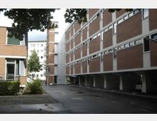 Mehrstöckige Wohngebäude aus rotem Backstein mit weißen Fensterrahmen, die einen Innenhof mit gepflasterter Zufahrt und Parkplatz begrenzen. Im Hintergrund sind ein weiteres Wohngebäude und ein Baum zu sehen, während der Vordergrund teils von Baumkronen beschattet wird.