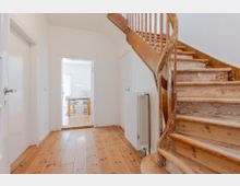 Heller Flur in einem Wohnhaus mit Holzdielenboden und einer geschwungenen Holztreppe mit Geländer an der rechten Seite. Durch eine Türöffnung ist ein angrenzender Raum mit Tisch und Stühlen sichtbar; an der Wand steht ein weißer Heizkörper.
