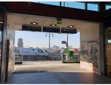 Blick aus einem überdachten Bereich eines Bahnhofs mit Graffiti an den Wänden auf die Warschauer Straße in Berlin, im Hintergrund geparkte Fahrräder, eine Straße mit einem Lieferwagen und die Skyline mit Hochhäusern. Oben befinden sich Wegweiser zu den Bahnsteigen und der Ausgangsbeschilderung.