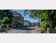 Straße in einem ländlichen Wohngebiet mit Einfamilienhäusern, flankiert von Bäumen und Zäunen. Links steht ein zweigeschossiges Haus mit rotem Ziegeldach, und die Straße führt an mehreren weiteren Häusern entlang in einen bewaldeten Bereich.