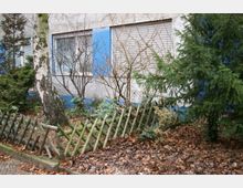Wohnhausfassade in grauer Putzoptik mit blauen Fensterrahmen und geschlossenen Jalousien, umgeben von einem kleinen Garten mit dünnen Sträuchern, einem Birkenbaum und immergrünen Nadelbäumen. Im Vordergrund ein schräger Holzzaun, Laub bedeckt den Boden.
