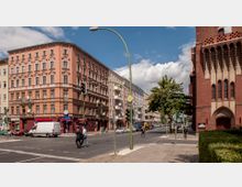 Straßenszene in einer Stadt mit einem mehrstöckigen, rosa Wohn- und Geschäftshaus mit aufwändiger Fassade links und einer roten Backsteinkirche mit hohen Fenstern und Spitzbogenportal rechts. In der Mitte befindet sich eine Kreuzung mit Verkehrsampeln, Fußgängern und parkenden Autos.