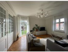 Wohnzimmer mit Parkettboden, großem Fenster und Glastüren, die auf einen Garten mit grüner Rasenfläche führen. Der Raum ist mit einem Sofa, einem Teppich, einem Lowboard mit TV und Pflanzen dekoriert; ein Kronleuchter hängt von der Decke.