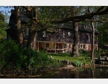 Zweistöckiges Haus mit dunkler Fassade und gemauertem Erdgeschoss, umgeben von Bäumen und einem Garten mit Sitzgelegenheiten. Im Vordergrund fließt ein kleiner Wasserlauf, ergänzt durch Ufervegetation und Holzpfosten.
