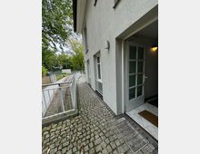 Gebäudeeingang mit offener weißer Tür, umgeben von hellgrauer Putzfassade. Der Weg vor dem Gebäude ist aus Kopfsteinpflaster und führt entlang eines Geländers; im Hintergrund sind Bäume und ein Zaun zu sehen.