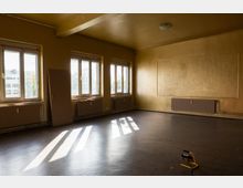 Leerer, renovierungsbedürftiger Innenraum mit gelblichen Wänden, drei großen Fenstern und Heizkörpern darunter; Sonnenlicht wirft lange Schatten auf den dunklen Boden. An der rechten Wand befindet sich eine große rechteckige Wandfläche, daneben lehnt eine Tür an der Wand.