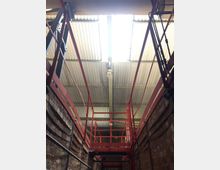 Industriehalle mit Wellblechdach, hellem Oberlicht und roten Metallkonstruktionen wie Geländern und Plattformen. Die Wände bestehen aus roten Ziegelsteinen, und es sind elektrische Leitungen sichtbar.
