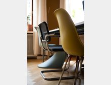 Ein moderner Innenraum mit verschiedenen Designerstühlen an einem Tisch, darunter ein gelber Kunststoffstuhl, ein schwarzer Stuhl mit Metallrahmen und ein hellblauer Freischwinger. Im Hintergrund sind ein Fenster mit Gardinen, eine Heizung und Zimmerpflanzen sichtbar.