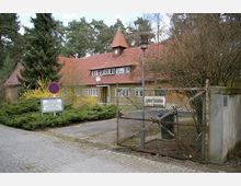 Eingezäuntes Gebäude mit gelbem Putz und roten Dachziegeln in ländlicher Umgebung, umgeben von Bäumen und Büschen. Im Vordergrund befinden sich ein Metallzaun, ein Tor sowie ein Schild mit Informationen und ein Parkverbotschild.