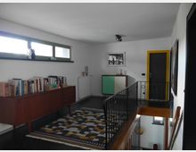 Ein Innenraum mit modernem Design, bestehend aus einer weißen Wand mit einem kleinen horizontalen Fenster oben, einer hölzernen Anrichte mit Büchern, einem Teppich mit geometrischem Muster und einer gelb umrahmten Tür im Hintergrund. Auf der rechten Seite ist ein Metallgeländer, das zu einer Treppe gehört.