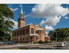 Backstein-Kirche im neoromanischen Stil mit grünem Turmdach und einer kupferfarbenen Kuppel, umgeben von Bäumen und geparkten Autos, vor einem blauen Himmel mit weißen Wolken. Im Vordergrund verläuft eine Straße mit einem parkenden Auto.