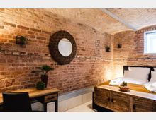 Gemütliches Schlafzimmer mit unverputzten Ziegelwänden, einem gemusterten Rundspiegel, einem Holztisch mit Pflanzen und einem Bett mit weißer Bettwäsche und Deko-Elementen. Ein kleines Fenster bringt Tageslicht in den Raum, der eine warme und rustikale Atmosphäre vermittelt.