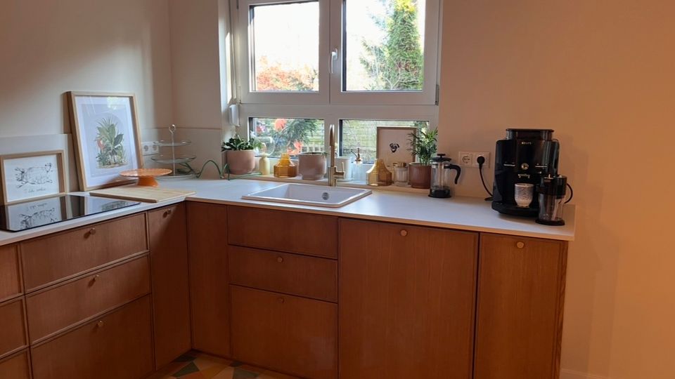 Küche mit Holzunterschränken, einer weißen Arbeitsplatte und einem Spülbecken unter einem Doppelfenster. Auf der Arbeitsplatte stehen Pflanzen, Küchenutensilien sowie eine Kaffeemaschine; der Boden ist mit geometrisch gemusterten Fliesen gestaltet.