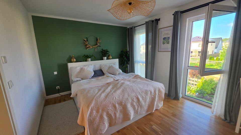 Modernes Schlafzimmer mit Doppelbett vor einer dunkelgrünen Akzentwand, hellen Betttextilien und Holzfußboden. Rechts sorgen große bodentiefe Fenster mit grauen Vorhängen und geöffnetem Flügel für viel Tageslicht und Blick ins Wohngebiet mit Garten.