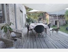 Holzterrasse mit einem Esstisch, der mit einer gemusterten Tischdecke bedeckt ist, umgeben von schwarzen Stühlen und Korbsesseln; darüber ein Sonnensegel und ein Sonnenschirm. Im Hintergrund steht ein Hund, umgeben von einer Ziegelmauer und einem begrünten Garten.