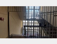 Treppenhaus mit einfachem Geländer aus Metall und mit Drahtgitter abgesichert. Im Hintergrund befinden sich große Fenster, die natürliches Licht hereinlassen; an der Wand ein Feuermelder und eine Steckdose.