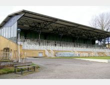 Historische Tribüne mit überdachtem Dach und offener Stahlkonstruktion, umgeben von Ziegelsteinmauern und weißen Geländern. Im Vordergrund befindet sich ein gepflasterter Hof mit einer Parkbank und Rasenflächen.