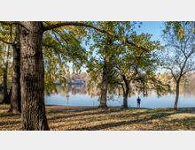 Grünanlage mit Bäumen am Ufer eines Sees, im Hintergrund ein Gebäude am gegenüberliegenden Ufer. Eine Person steht nahe dem Wasser, umgeben von herbstlichem Laub auf dem Boden.
