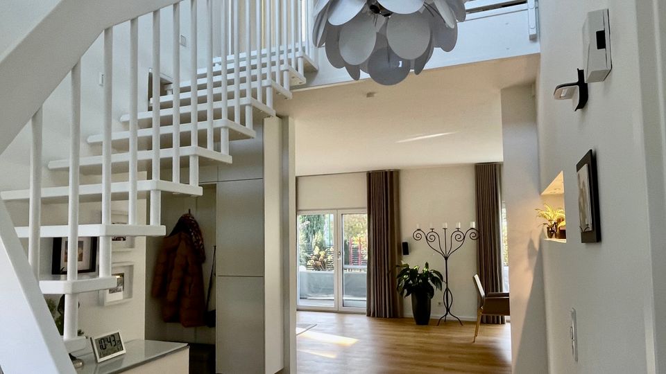 Heller, moderner Wohnungsflur mit offener weißer Treppe und Geländer, Holzfußboden und einer großen, blütenförmigen Pendelleuchte im Luftraum. Im Hintergrund führt eine breite Öffnung in einen Wohnbereich mit großen Fenstern und Vorhängen, Pflanzen und einem schlanken Kerzenständer.