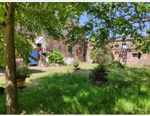 Grüner Innenhof eines alten Backsteingebäudes mit gemähtem Rasen, Bäumen und Sträuchern. Im Hintergrund ist ein Gebäude mit blauer Holztür und einer kleinen Treppe erkennbar, auf der Personen sitzen.