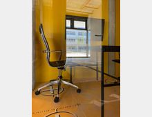 Ein moderner, kleiner Büroraum mit gelben Wänden und Glaswand, ausgestattet mit einem schwarzen Bürostuhl auf Rollen und einem schwarzen Schreibtisch. Im Hintergrund ist ein Fenster mit Blick auf ein Bürogebäude und einen Außenbereich mit Bäumen und Sitzgelegenheiten zu sehen.