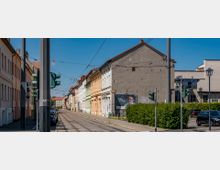 Straße mit Straßenbahnschienen und historischen Wohnhäusern in hellen Farben auf der linken Seite, flankiert von einer grauen Hausfassade rechts. Im Vordergrund Ampeln, geparkte Autos und ein Werbeplakat, im Hintergrund ein blauer Himmel.