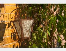 Eine rustikale, metallene Außenlaterne mit dekorativen Verzierungen hängt vor einer gelben Backsteinwand. Im Hintergrund sind grüne Kletterpflanzen zu sehen, die das natürliche und warme Ambiente betonen.