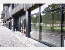 Moderne Gebäudefassade mit großen Glasfronten und dunklen Metallrahmen, angrenzend an eine gepflasterte Gehwegfläche. Im Vordergrund sind ein Mietfahrrad und E-Scooter aufgestellt, während im Hintergrund Bäume und eine grüne Parklandschaft sichtbar sind.