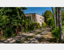 Einfamilienhaus mit flachem Dach und verputzter Fassade, umgeben von dichtem Grün und großen Bäumen. Ein schmaler, unbefestigter Weg führt entlang eines niedrigen Gitters mit Pflanzenbewuchs zur Eingangstür.