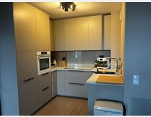 Moderne, kompakte Einbauküche in L-Form mit hellen, grifflosen Schränken und integrierten Geräten wie Backofen und Kochfeld. Auf der Arbeitsplatte stehen Spüle, Kaffeemaschine und Küchenutensilien; der Boden ist in Holzoptik und die Beleuchtung erfolgt über eine Deckenleuchte.