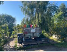 Ein Gemeinschaftsgarten mit einem zentralen Bewässerungsbereich, bestehend aus einem großen Wassertank, Gießkannen in verschiedenen Farben und Recycling-Behältern. Umgeben von üppigem Grün, Bäumen und Blumenbeeten, mit einem schmalen Pfad im Vordergrund.