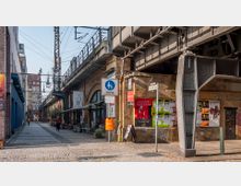 Eine gepflasterte Straße unter einer erhöhten Eisenbahntrasse aus Stahl, flankiert von Backsteingebäuden mit Arkaden. An den Gebäuden befinden sich Geschäfte und Cafés, sowie bunte Plakate; auf der Straße stehen Straßenschilder und ein orangefarbener Mülleimer.