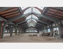 Geräumige, leerstehende Halle mit sichtbaren Gitterträgern aus Metall und Holzelementen, einem hohen Glasdach und großen Fenstern an den Seiten; Baumaterialien sind unordentlich in einem Bereich gestapelt.