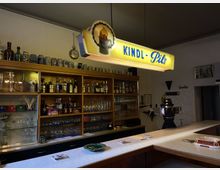 Ein klassischer Kneipen-Innenraum mit einer Bar und Regalen, die mit Gläsern, Spirituosenflaschen und Krügen gefüllt sind. Über der Theke hängt ein beleuchtetes Schild mit der Aufschrift "Kindl-Pils", und im Hintergrund ist eine Zapfanlage sichtbar.