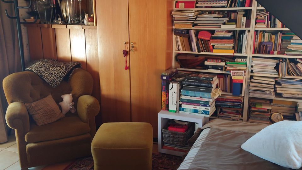 Gemütlicher Wohnraum mit Holzverkleidung, einem großen Bücherregal voller Bücher, einem gelben Sessel mit passendem Fußhocker und einem Bett im Vordergrund. Eine schwarze Treppe führt nach oben, und ein bunter Teppich bedeckt den Boden.