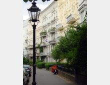 Straße mit historischen Wohnhäusern im Gründerzeitstil, verziert mit Balkonen und Stuckelementen; im Vordergrund steht eine Laterne, und rechts säumen Bäume und Büsche den Gehweg. Links parken Autos, und ein abgedeckter Motorroller steht am Straßenrand.