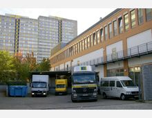 Hinterhof eines Gewerbegebäudes mit geparkten Lieferfahrzeugen, darunter Lastwagen von EDEKA und DHL. Im Hintergrund ein Hochhaus mit grauer Fassade und gelben Akzenten sowie Bäume mit herbstlichem Laub.