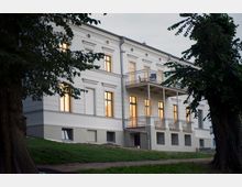 Ein zweistöckiges, historisches Gebäude mit weißer Fassade, verzierten Fensterrahmen und zwei Balkonen aus Metall. Im Vordergrund befinden sich Rasenflächen, ein Weg und große, dicht belaubte Bäume.